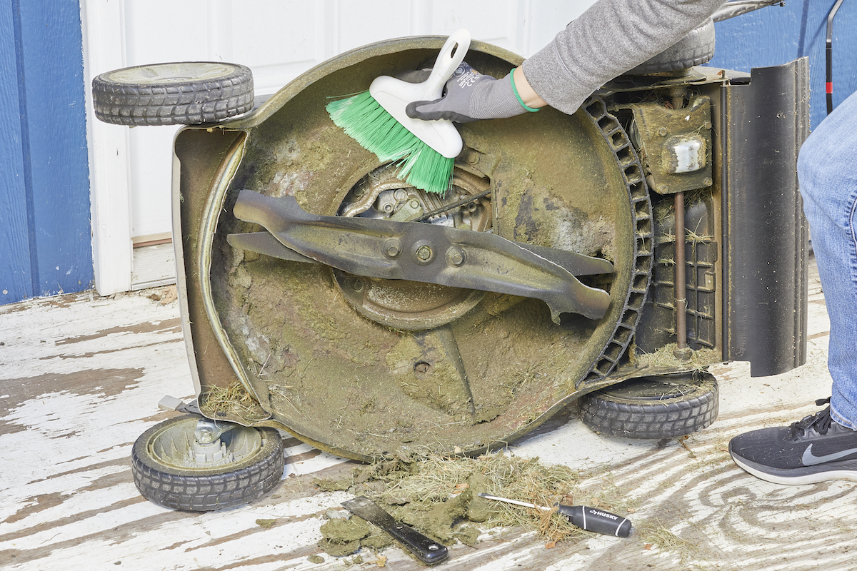Person uses a hand broom to clean underside deck of lawn mower.