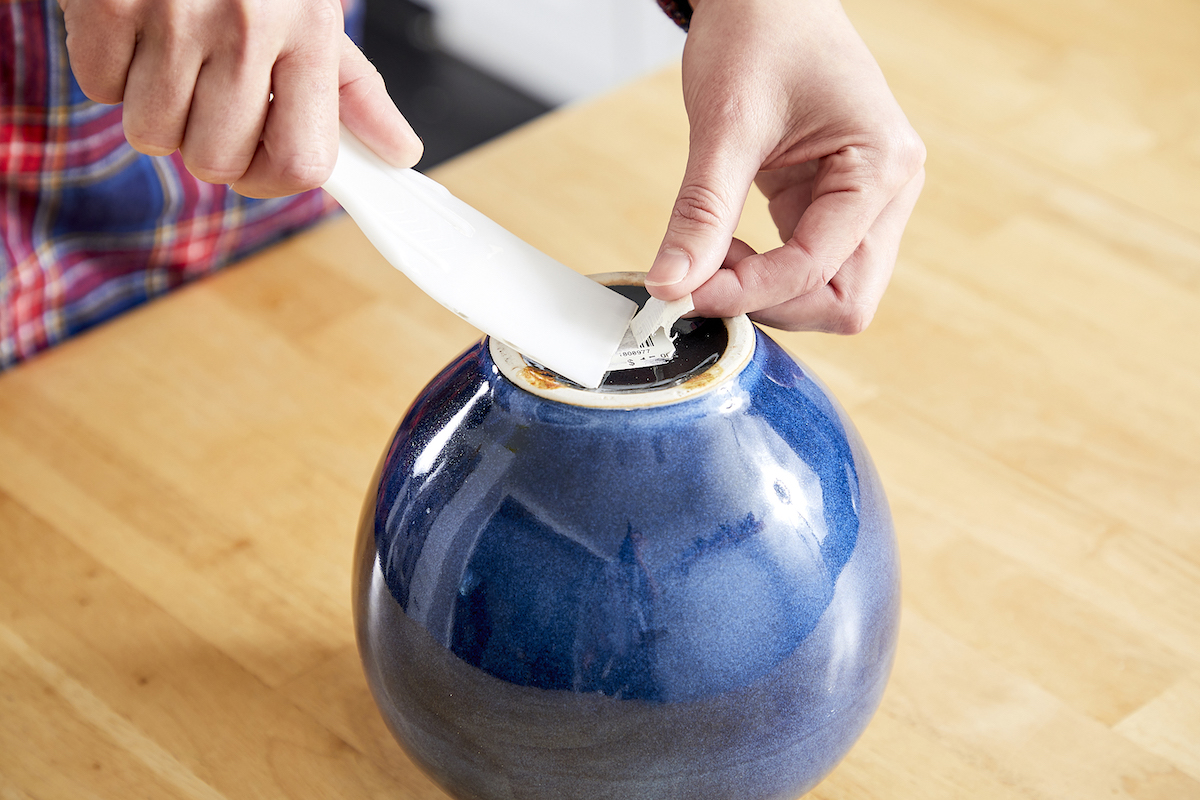Woman uses a putty knife to remove some of a sticker from the bottom of a jar.