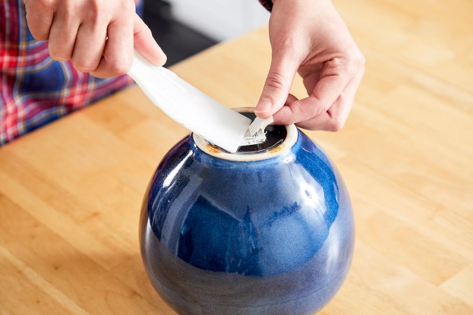 Woman uses a putty knife to remove some of a sticker from the bottom of a jar.