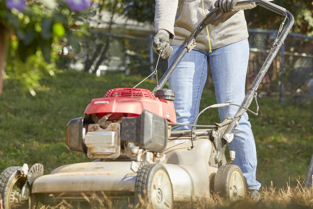 Woman wearing jeans pulls start cord on a red lawn mower.