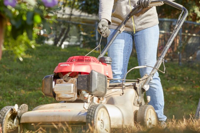 Woman wearing jeans pulls start cord on a red lawn mower.