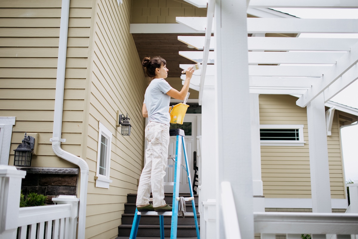 A woman wearing white paints a white deck with a gazebo while on a ladder.