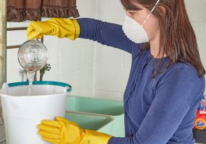 Woman wearing N95 mask pours liquid from measuring cup into large white bucket in a laundry room.