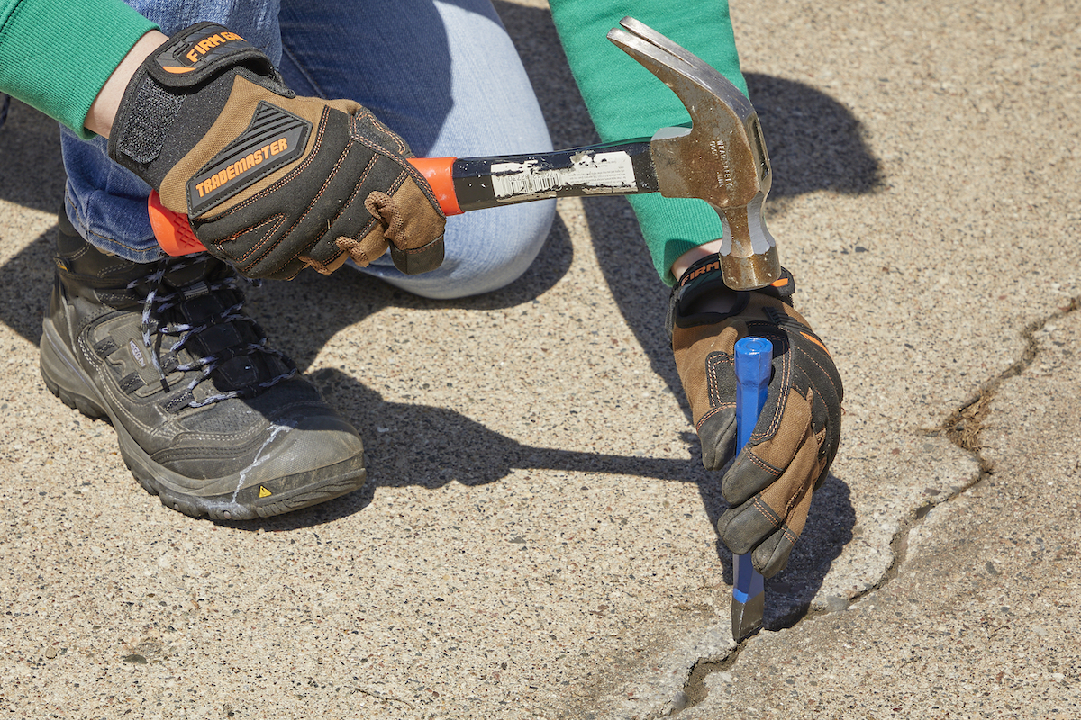 Woman uses a hammer and chisel to clean debris out of a crack in a concrete driveway.