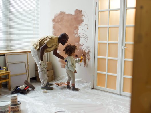 A father teaching his daughter how to use painting tools to paint a wall.