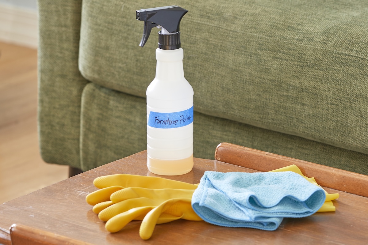 A spray bottled labeled "furniture polish" with painter's tape, along with a set of gloves and a microfiber cloth, on a coffee table in front of a couch.