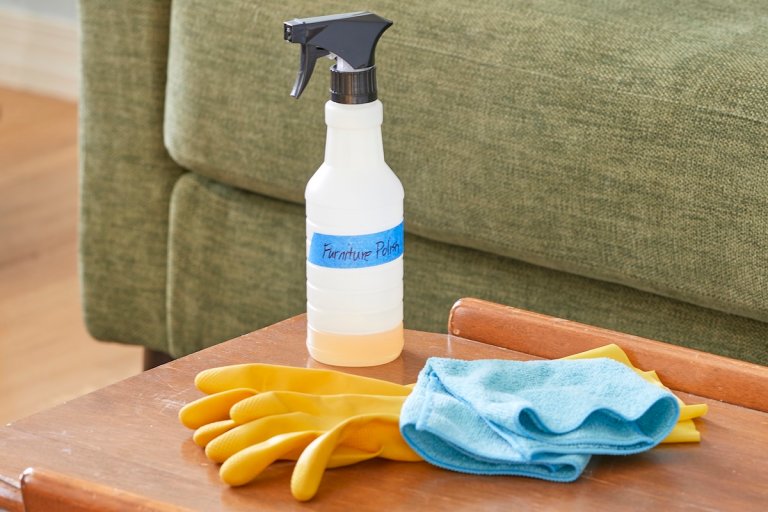 A spray bottled labeled "furniture polish" with painter's tape, along with a set of gloves and a microfiber cloth, on a coffee table in front of a couch.
