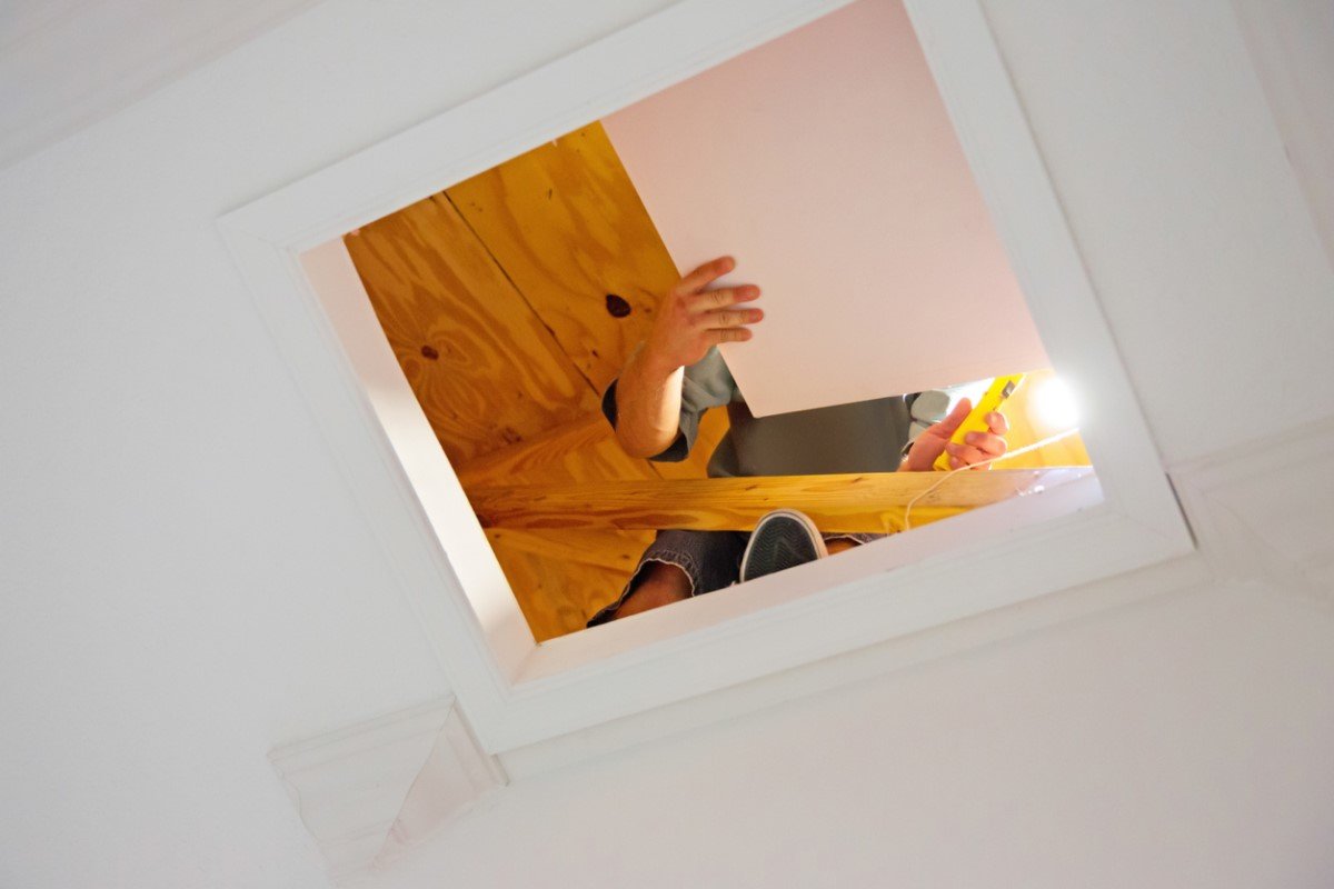View of person in attic removing access panel in ceiling to attic.