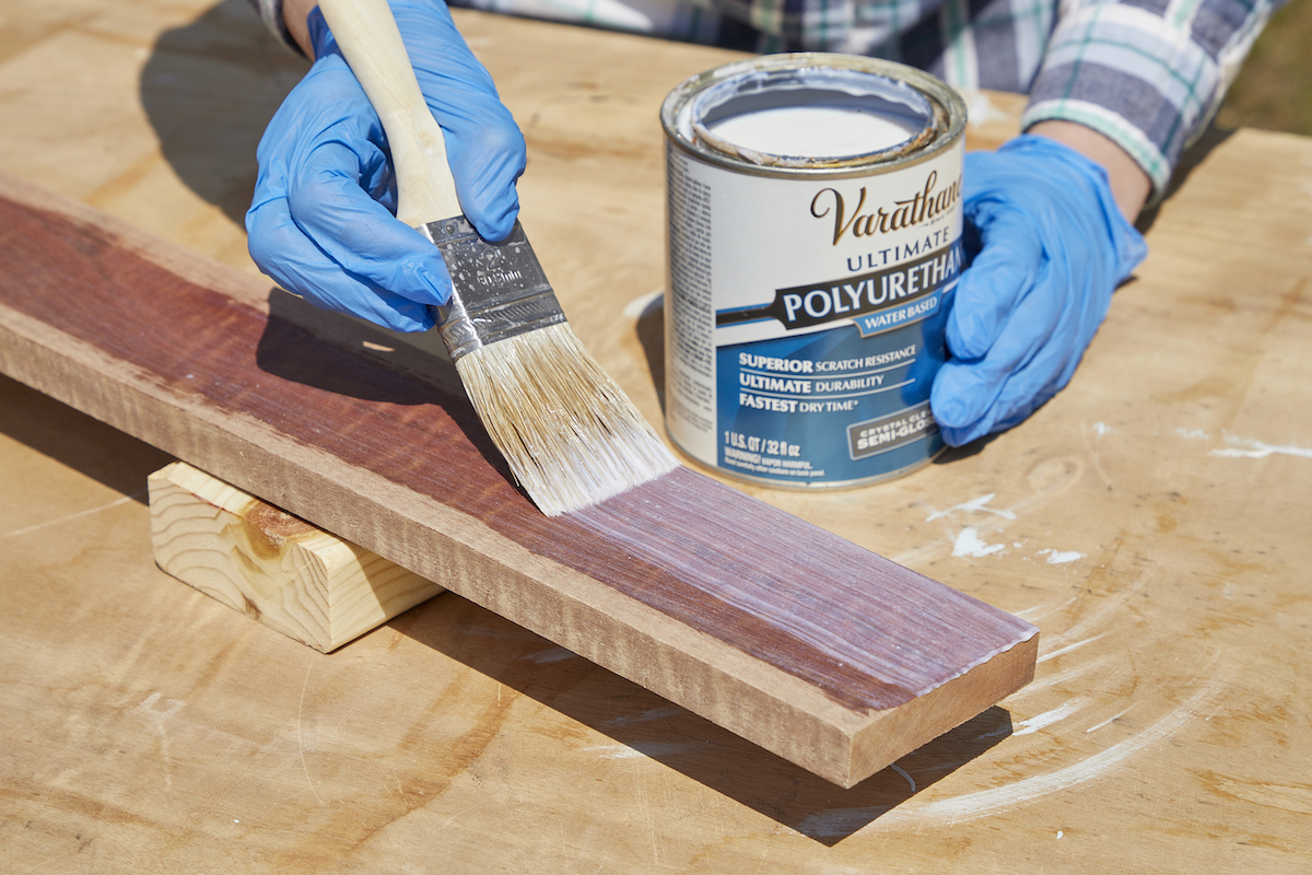 Woman uses a bristle paintbrush to apply polyurethane to a piece of wood.