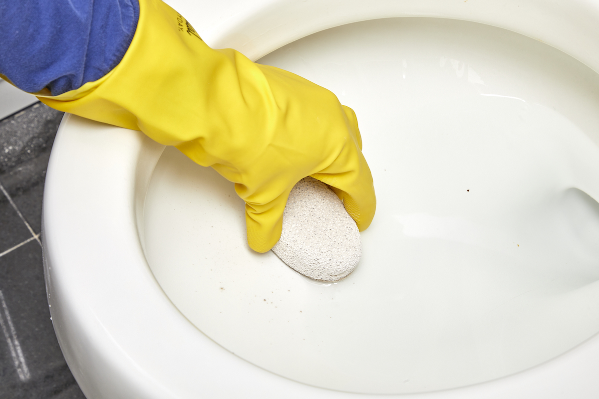 Woman wearing yellow rubber glove cleaning interior of toilet with a pumice stone.