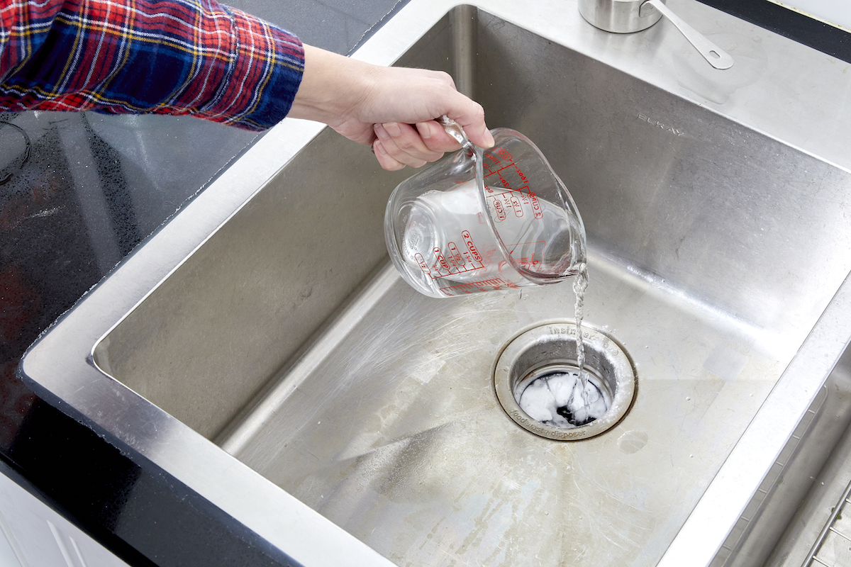 Woman pours measuring cup of vinegar into a garbage disposal with baking soda in the opening.