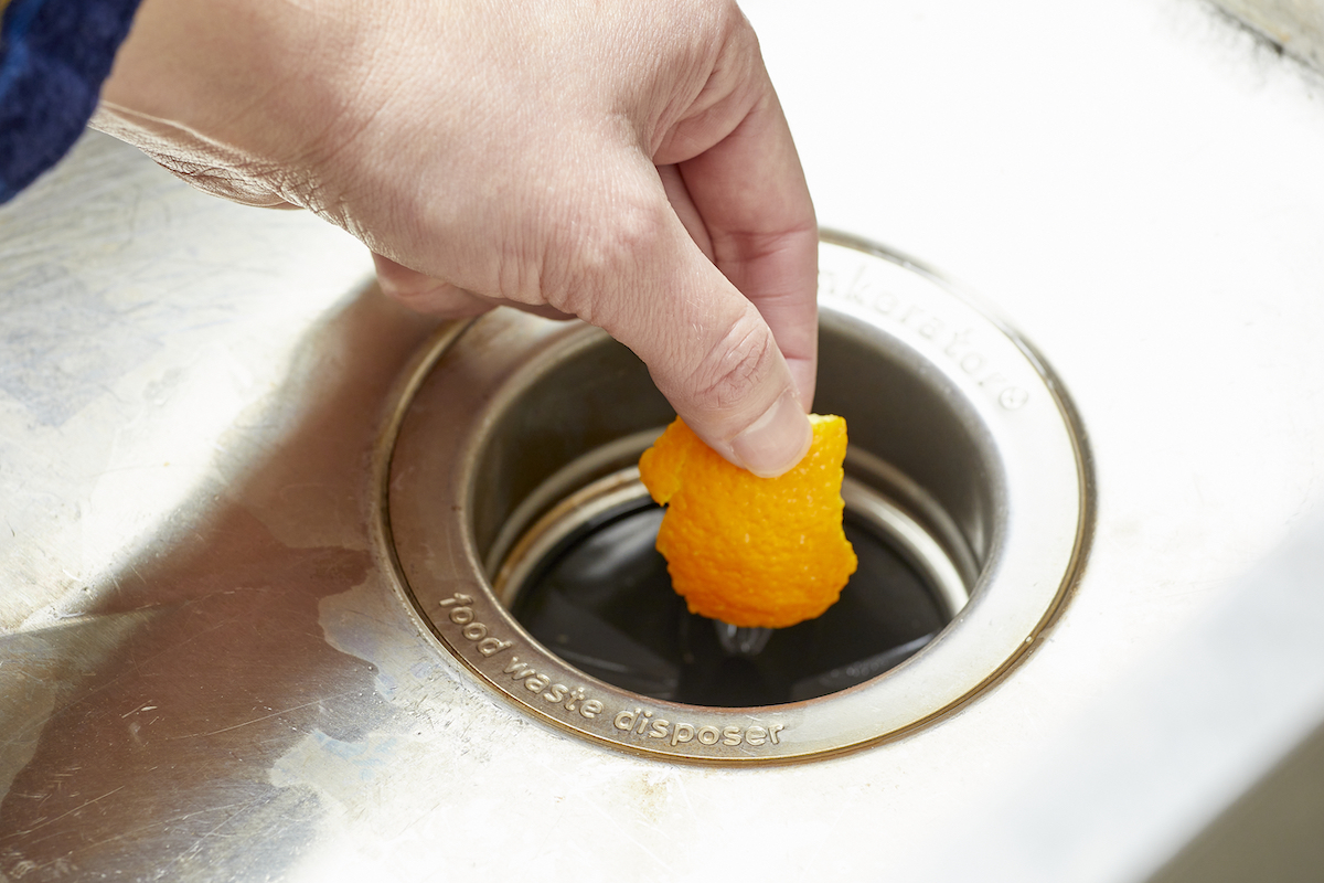 Woman holds orange peel over a garbage disposal.