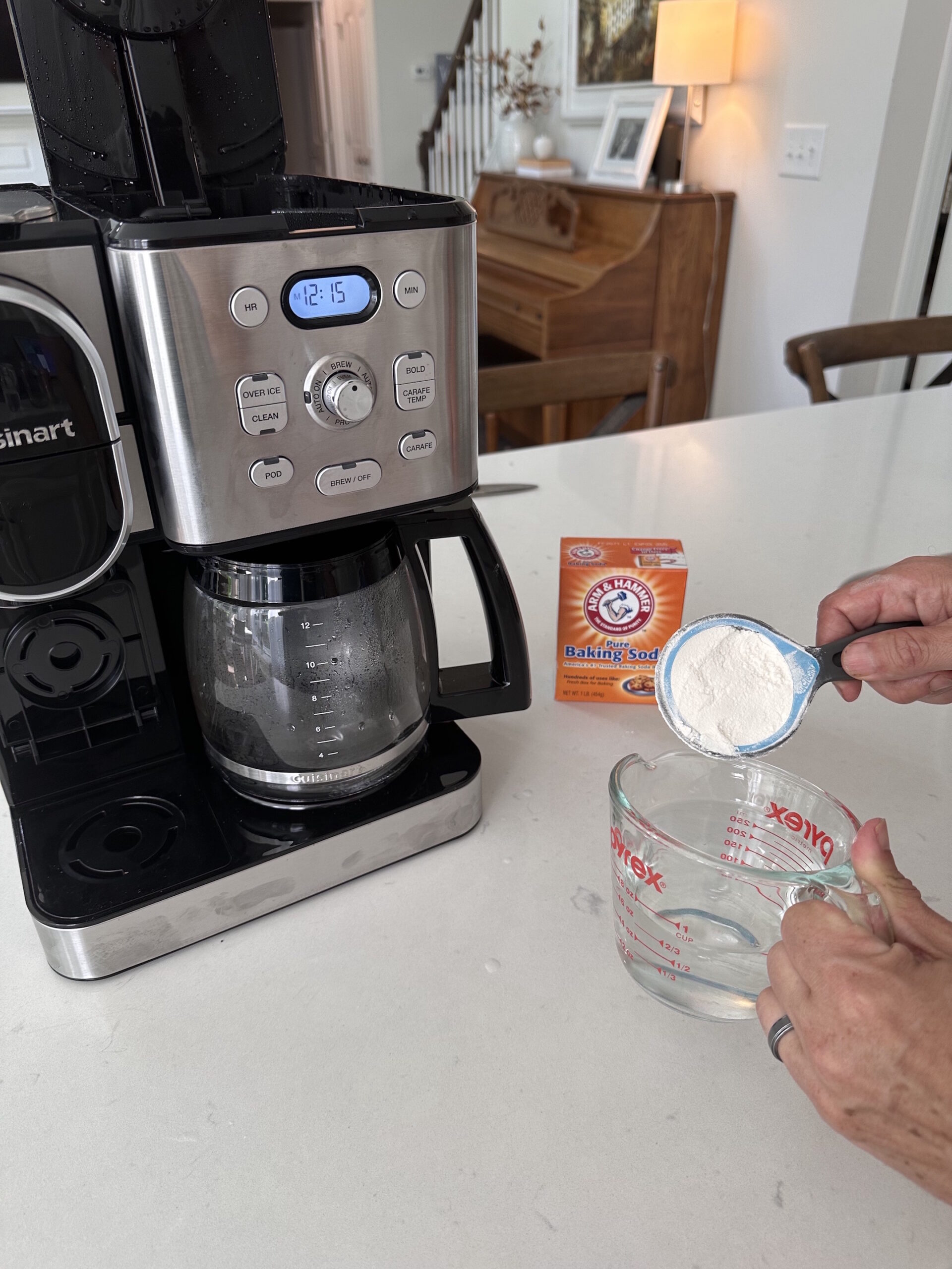 dropping a scoop of baking soda into a measuring cup with coffee maker on kitchen counter