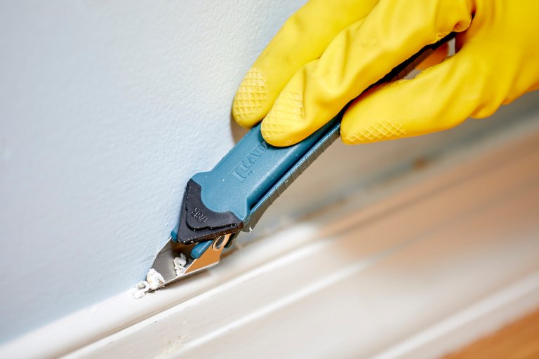 Woman wearing yellow gloves uses a caulk remover tool to remove caulk from atop a baseboard.