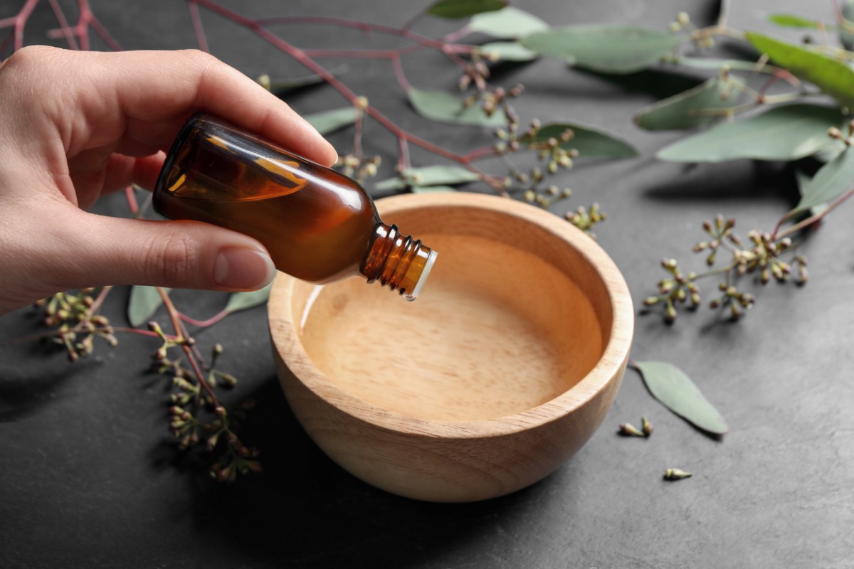 a woman's hand holding a small bottle of essential oils and dropping a few drops into a wooden bowl of water
