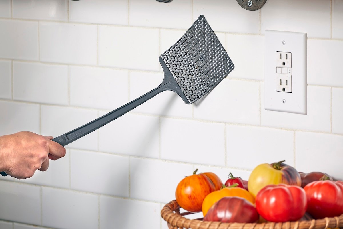 Closeup of woman's hand holding a fly swatter against a kitchen backsplash to kill a fly.