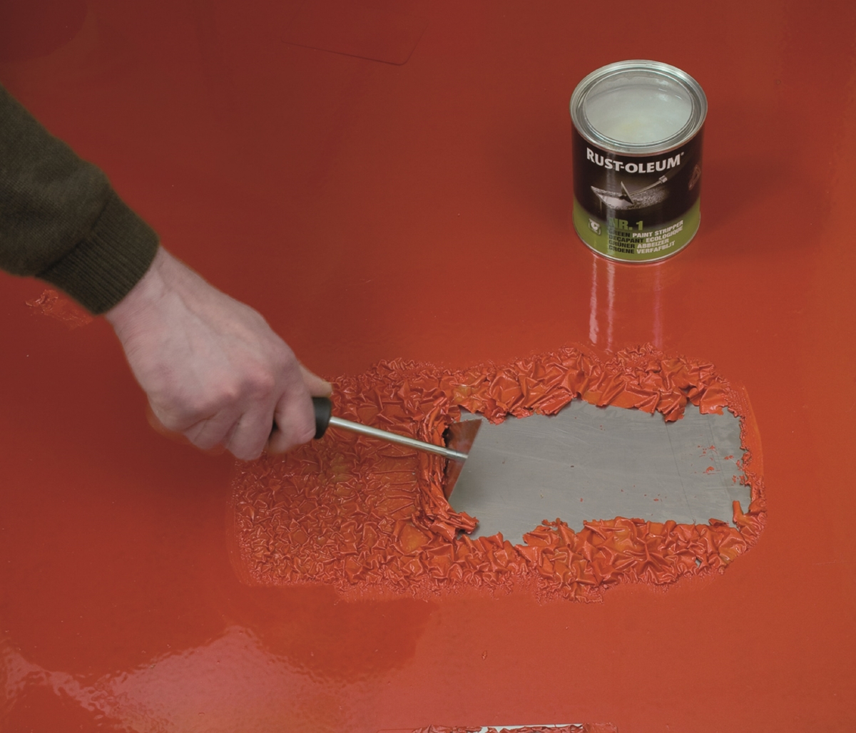 A hand using a tool to strip red paint off metal.