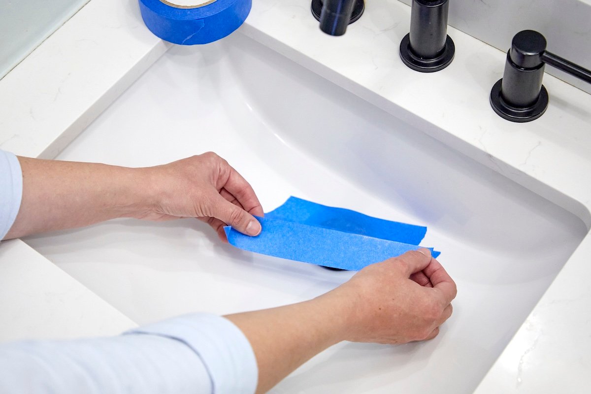 Woman rips a piece of blue painter's tape and prepares to place it over a bathroom sink drain.