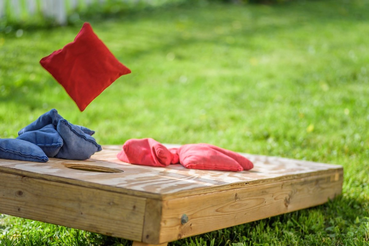 A wood cornhole board in the yard, with blue and red cornhole bags flying above, and sitting atop, the wood board.