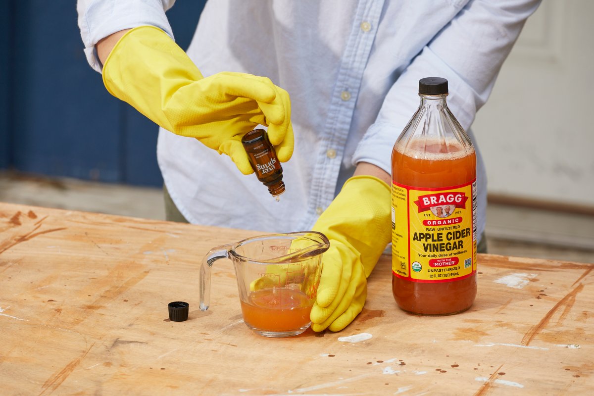 Woman squeezes essential oils into a measuring cup filled with apple cider vinegar, a bottle of apple cider vinegar nearby.