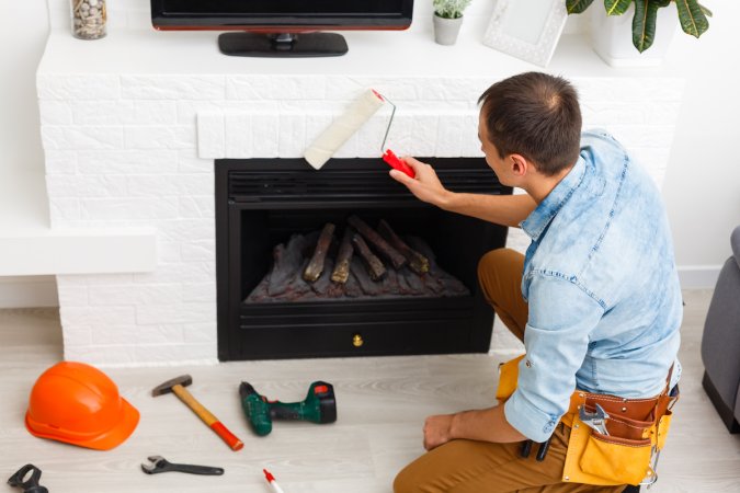 Handyman kneels in front of a white brick fireplace, painting it with a paint roller.