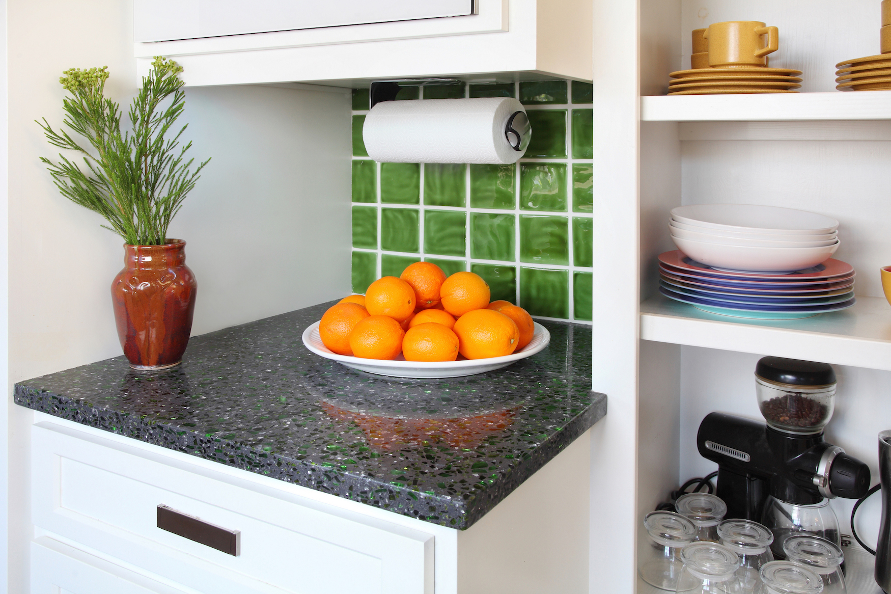 A small kitchen counter with a bowl of oranges next to an open shelving unit with mugs, plates, and appliances.
