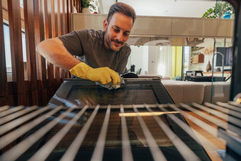 A smiling man wearing yellow gloves cleans the interior of an oven's window.