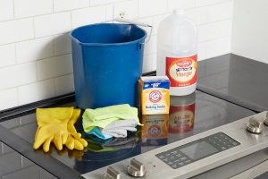 Blue bucket, vinegar, rubber gloves, and otherc cleaning materials on top of a glass cooktop.