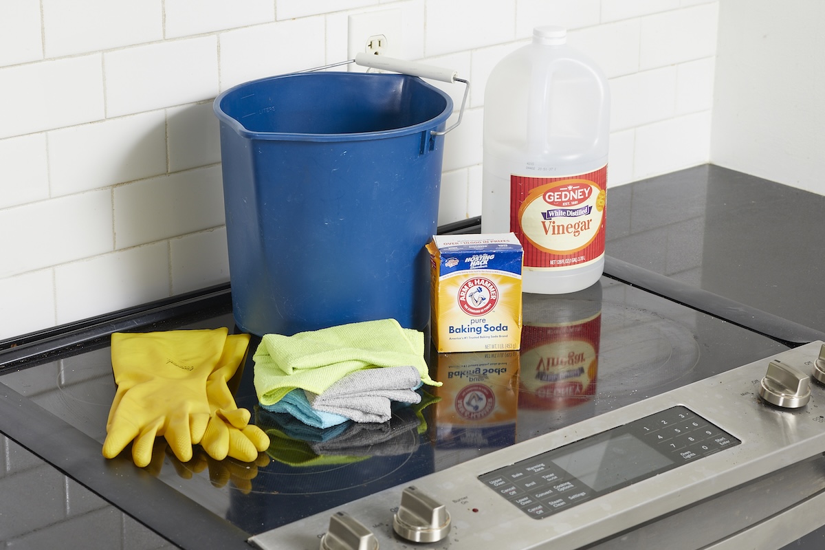 Blue bucket, vinegar, rubber gloves, and otherc cleaning materials on top of a glass cooktop.