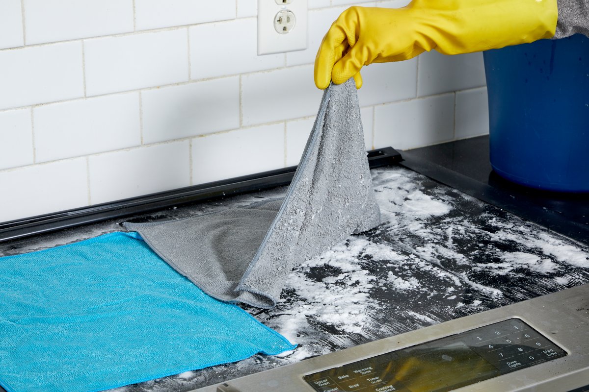 Person wearing yellow rubber gloves lifts damp microfiber cloths off baking soda mixture on glass cooktop.