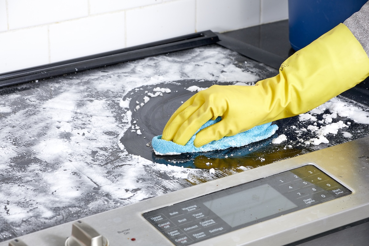 Person wearing rubber gloves scrubs glass cooktop with baking soda and blue microfiber cloth.