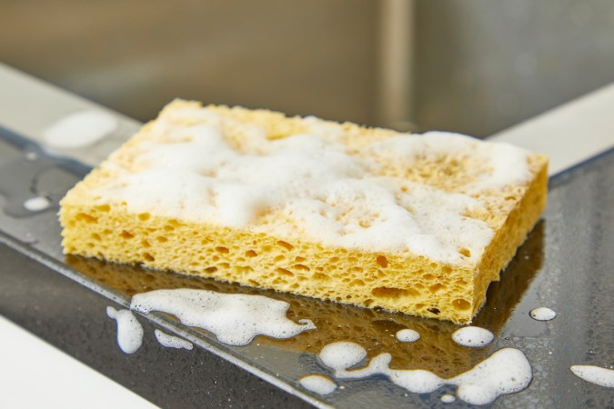 A soapy yellow dish sponge sits on the edge of a kitchen sink.