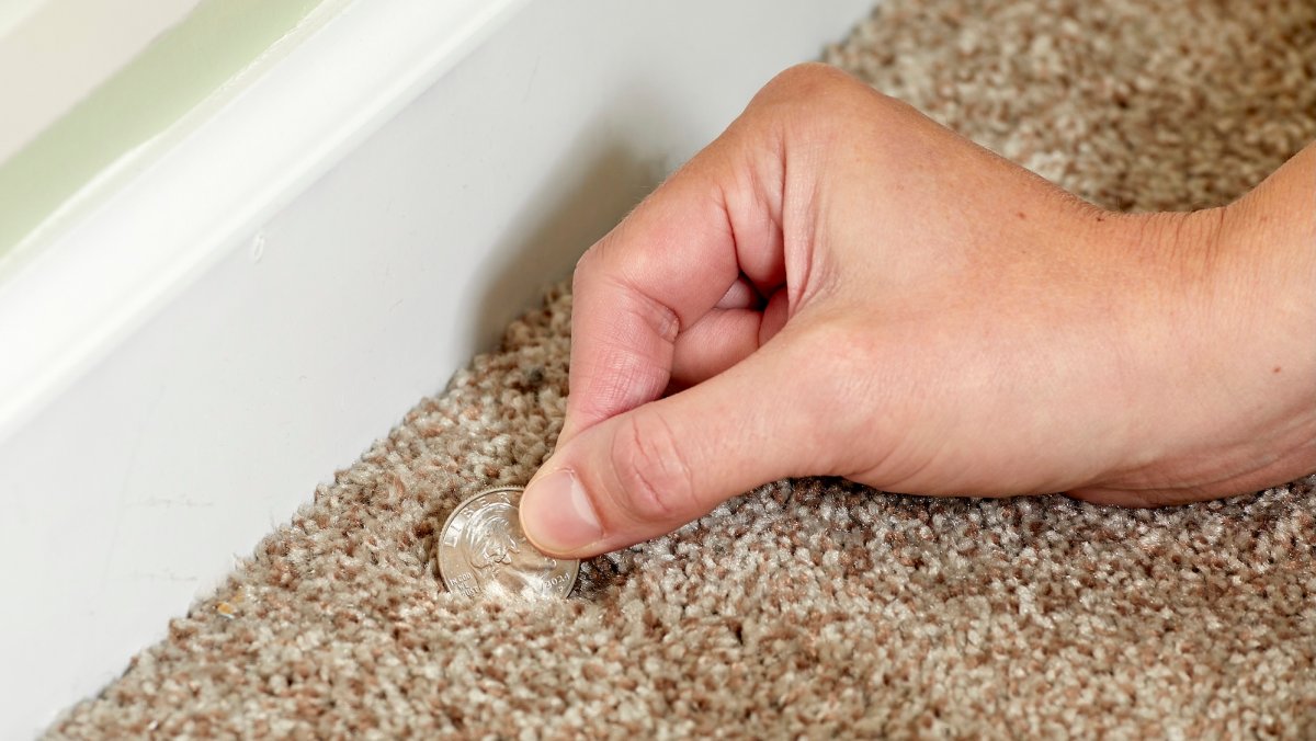 Woman uses a quarter to fluff a dent in a beige carpet.
