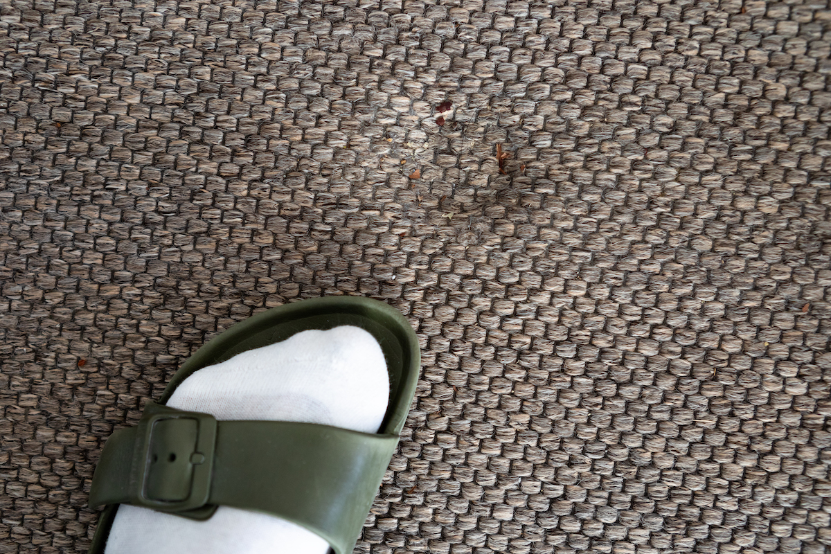 Person wearing a sandal with white socks standing on a brown carpet in front of an indentation in the carpet.