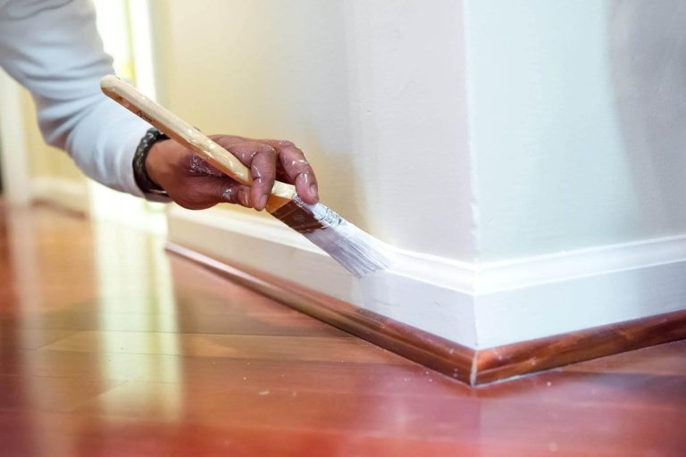 A closeup image of a man painting baseboards with an angled brush.