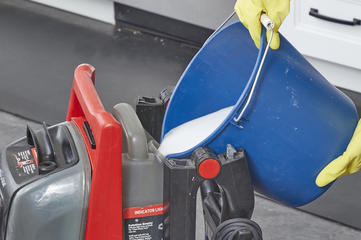 Person pours cleaning solution from blue bucket into a carpet cleaning machine's reservoir.