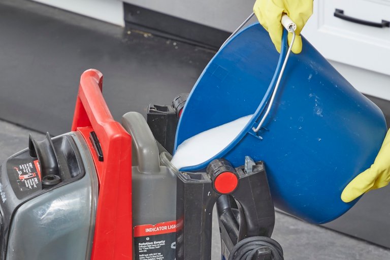 Person pours cleaning solution from blue bucket into a carpet cleaning machine's reservoir.