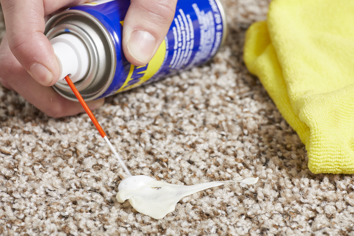 Person sprays WD-40 on a wad of gum on the carpet, a yellow cloth nearby.
