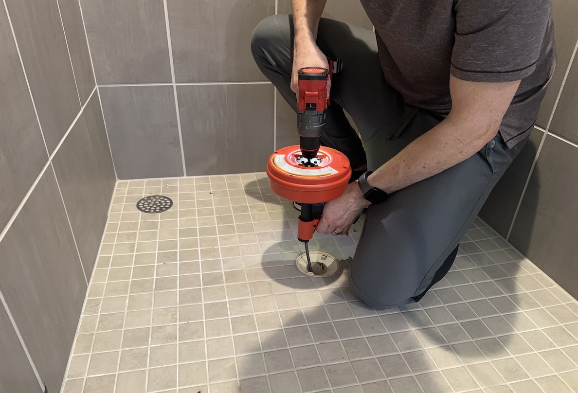 Man crouches in shower with a drain auger poised over the shower drain.