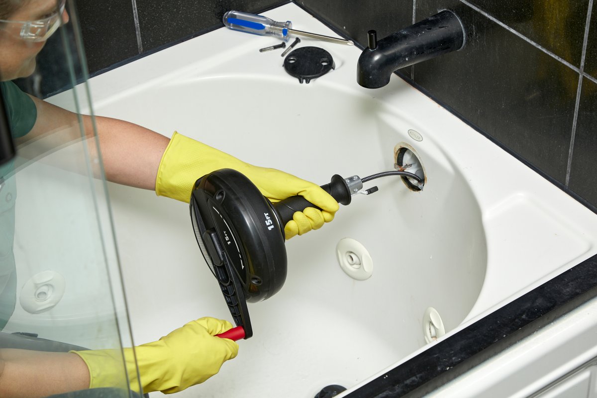 A woman wearing goggles and gloves twists a drain auger in a bathtub drain.