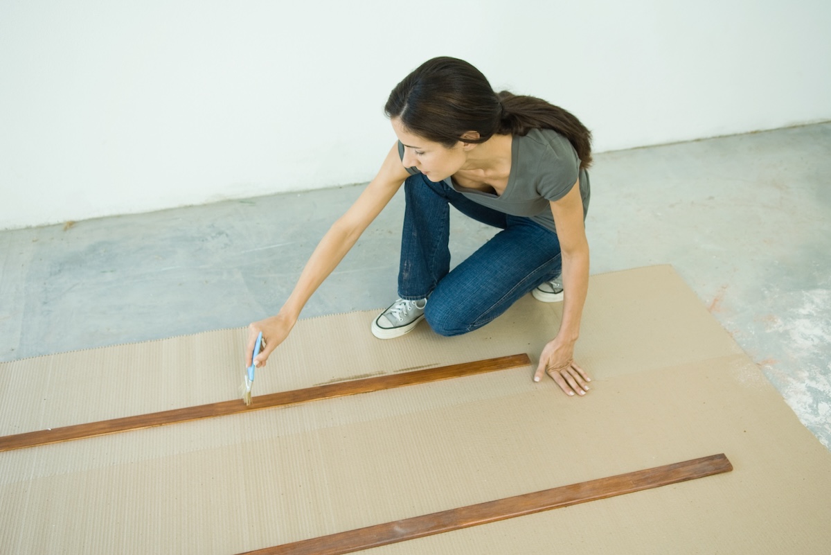 A woman paints baseboards that have been removed from the wall on a patch of cardboard.