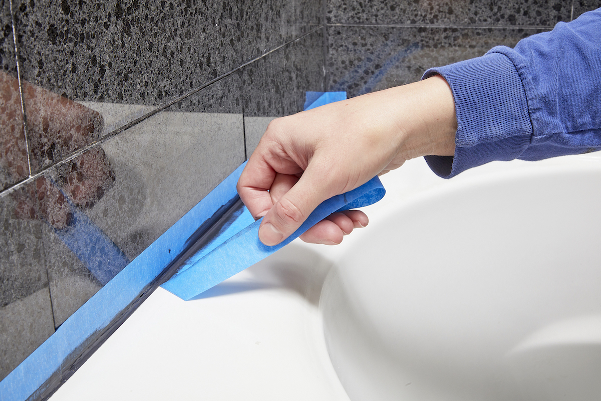 Woman peels painter's tape off tile and tub edge after caulking the joint.