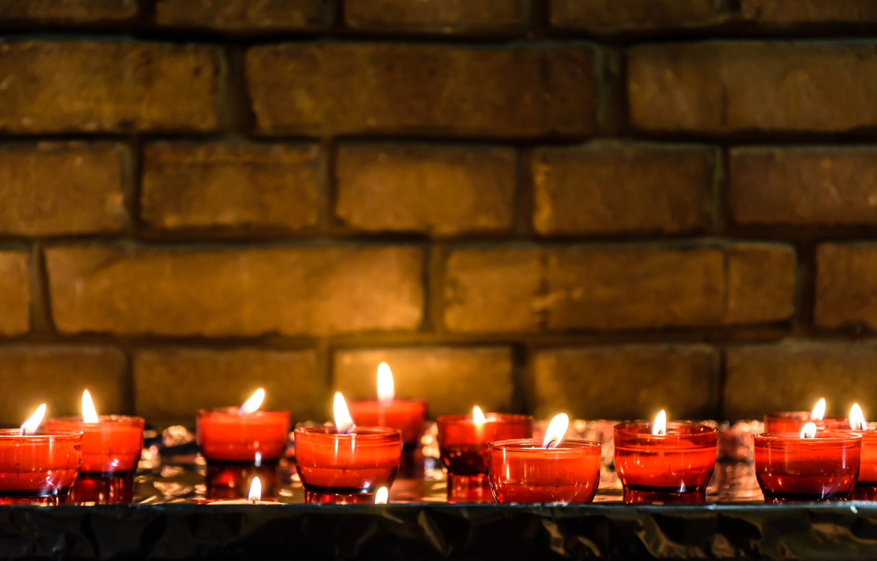 Small white votive candles lighted and placed in red glass holders on a table covered with aluminium foil in front of a brick wall.