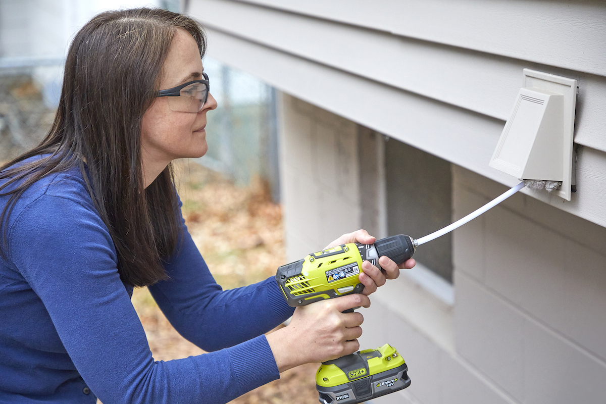 Woman uses a drill attachment to clean a dryer vent's exterior.