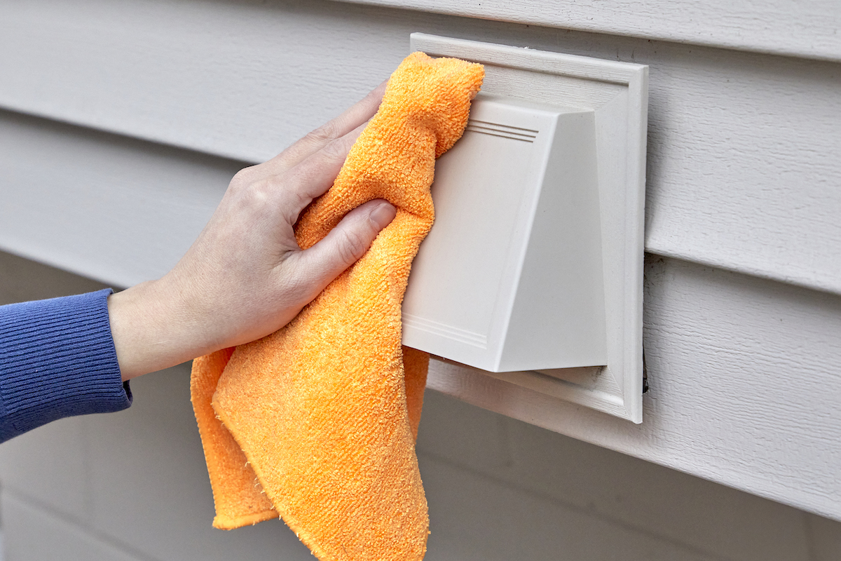 Woman wiping exterior dryer vent cover with orange microfiber cloth.