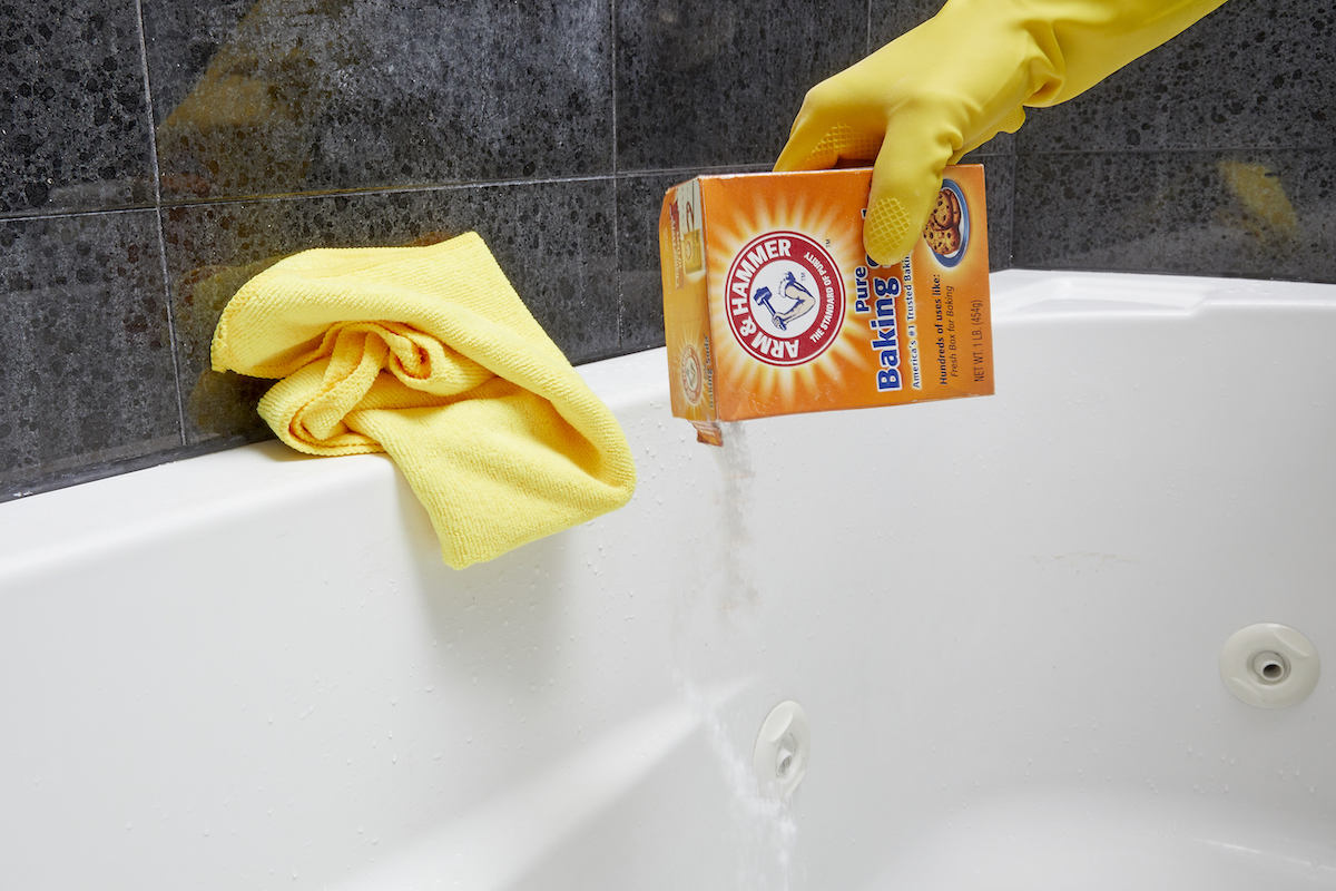 Woman wearing yellow rubber glove pours baking soda from a box into a jetted bathtub.