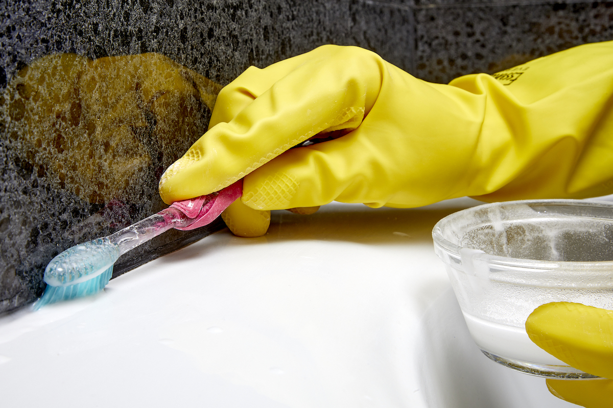 Woman wearing yellow rubber gloves scrubs the caulk around her jetted bathtub using a tootbrush.