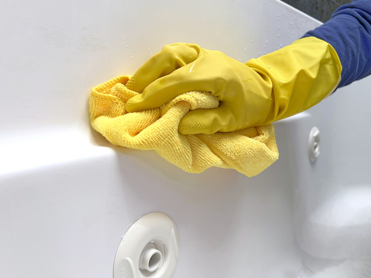 Woman wearing yellow rubber gloves wipes the inside of a jetted tub with a microfiber cloth.