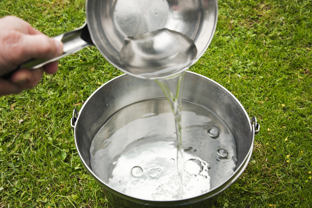Pouring fresh clear sparkling water from ladle into a bucket placed on green lawn in the garden.