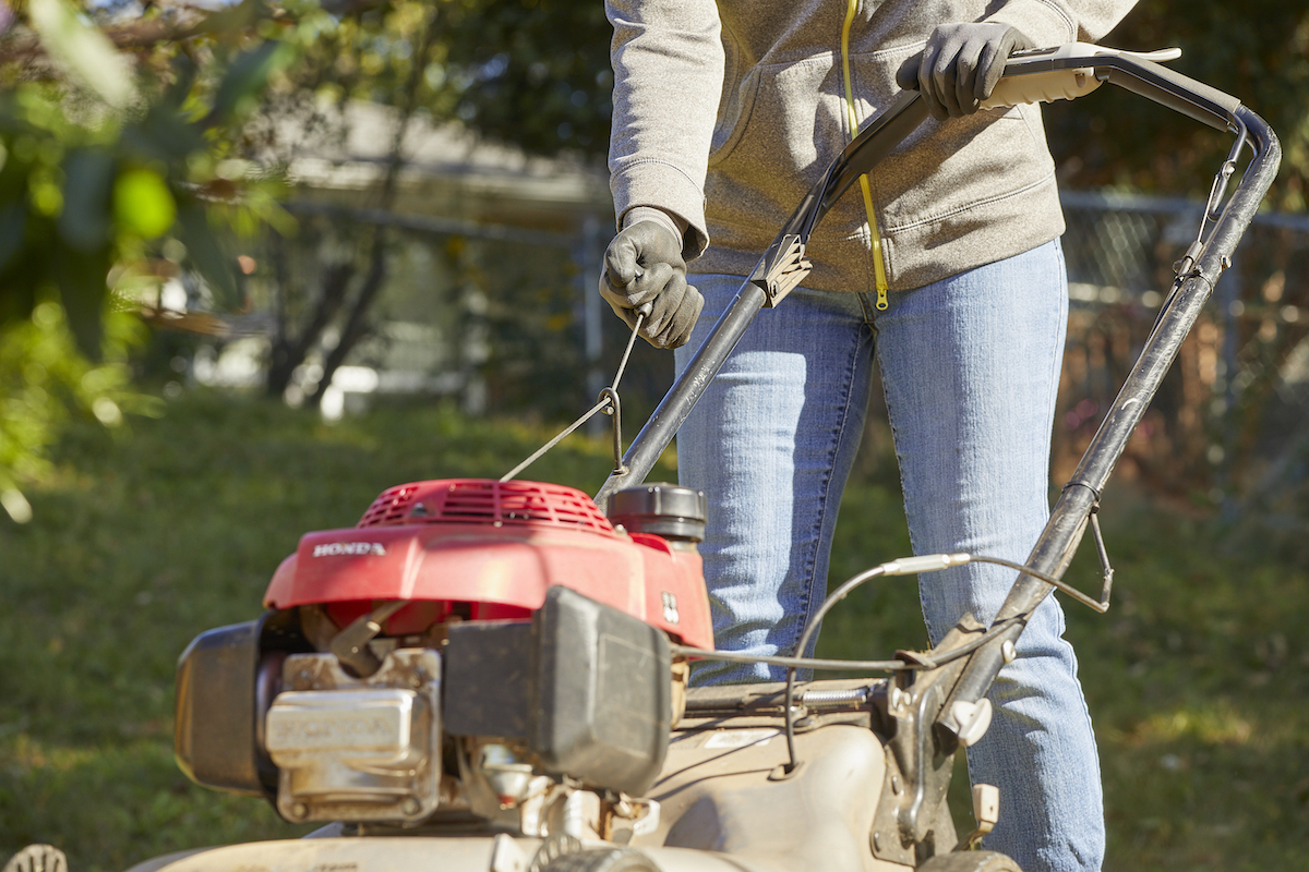 Woman wearing sweatshirt tries to start lawn mower in her yard.